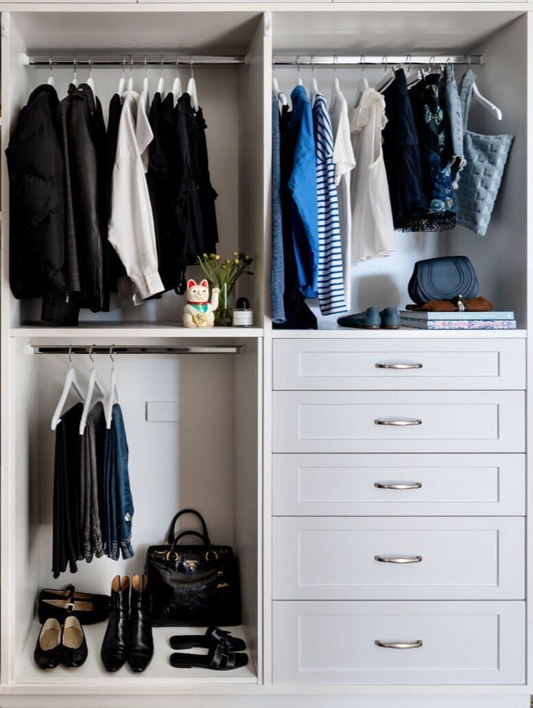 A wardrobe with black and blue clothing on a hanging rail, a handbag on the shelf above and white drawers below.