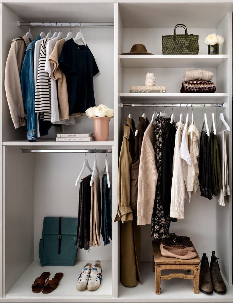 An open wardrobe with folded knitwear on the left shelves and a hanging rail of neutral clothing on the right.