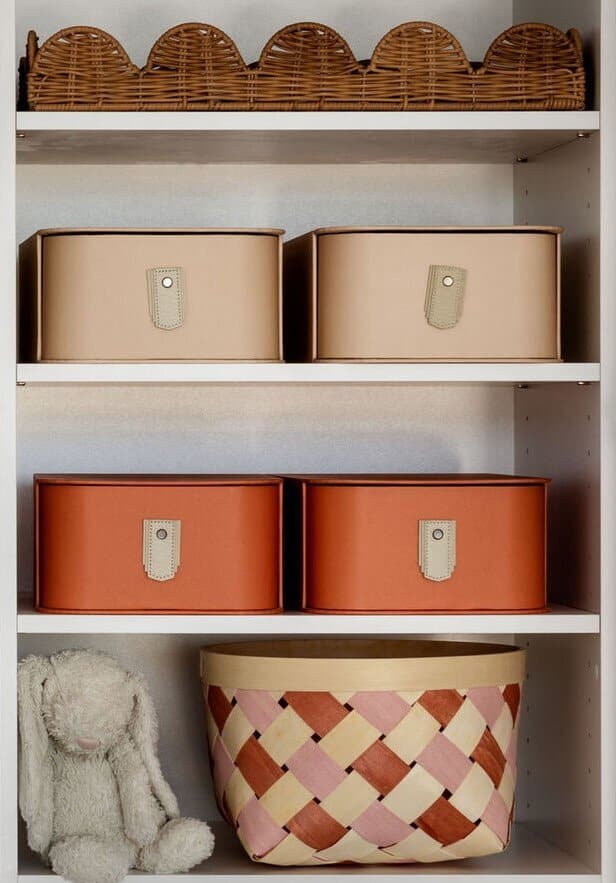 Stacked labelled storage boxes in tan and burnt orange on white shelving, with a patterned round hat box below.