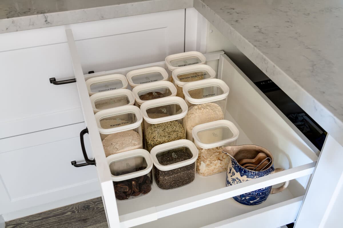 An open kitchen drawer fitted with white oval containers holding nuts and dried goods, beside two small blue patterned bowls.