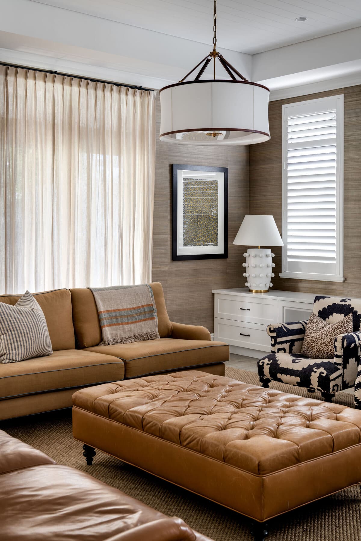 A living room with a tan leather sofa, buttoned leather ottoman, framed artwork and a table lamp against a grasscloth wall, with plantation shutters behind.