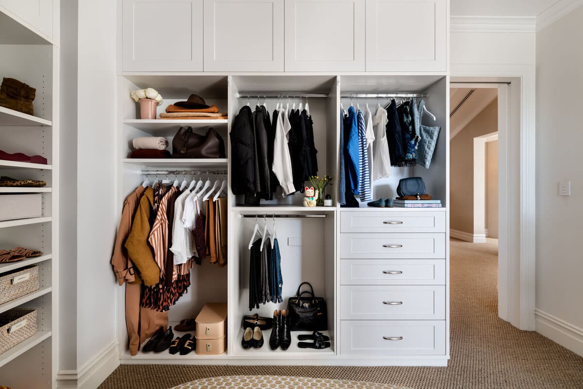 A built in wardrobe with hanging clothes, white drawers below and handbags on the upper shelf.