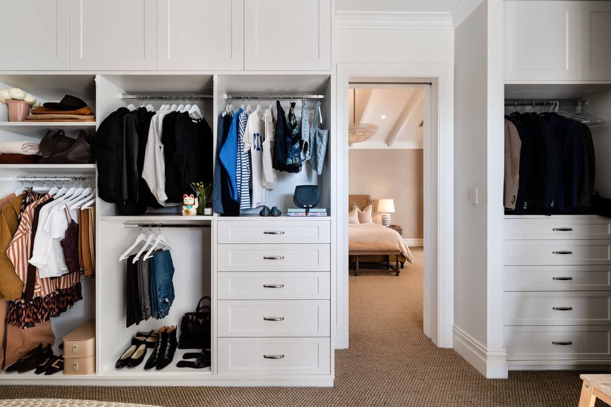 A wardrobe of hanging clothes and white drawers, opening through to a bedroom beyond.